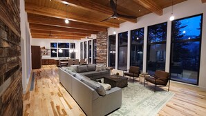 A modern living room with a sectional sofa, a coffee table, and a rug. There are large windows with a view of trees and a dining area with a table and chairs. - Amazing New 5 Bed Home next to the Columbia River. Sleeps 10 adults & 6 Kids (Stevenson)