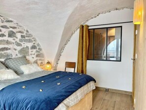 Ein Schlafzimmer mit Steinsandsteinwand, einem Bett mit blauer Decke, einem Nachttisch, einer Lampe und einem Fenster mit Vorhängen.