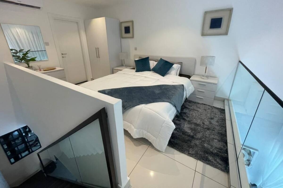 A modern hotel room with a large bed, a small desk, a chair, a nightstand, a lamp, a mirror, a window, and a door.
