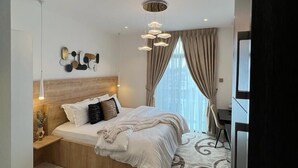 Un dormitorio moderno con cabecera de madera, una cama grande con ropa de cama blanca, una mesita de noche con lámpara, una silla y una alfombra estampada.