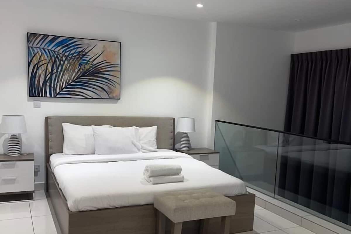 A modern hotel room with a large bed, a bedside table, a nightstand, a window with a view, and a painting on the wall.