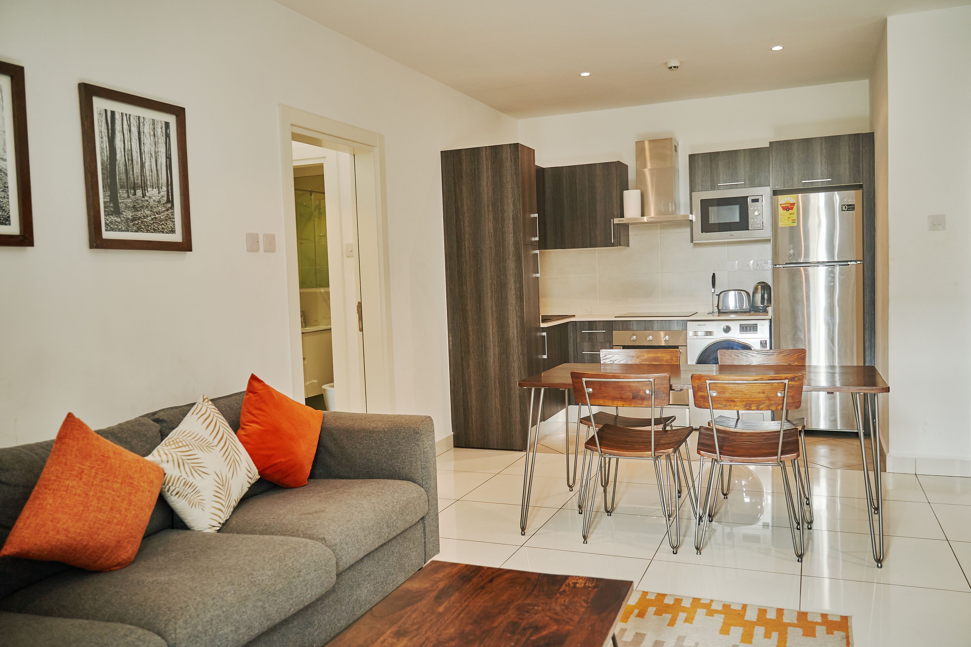 A modern living room with a grey sofa, orange and white cushions, and a wooden coffee table. A dining area with a wooden table and chairs is visible in the background.