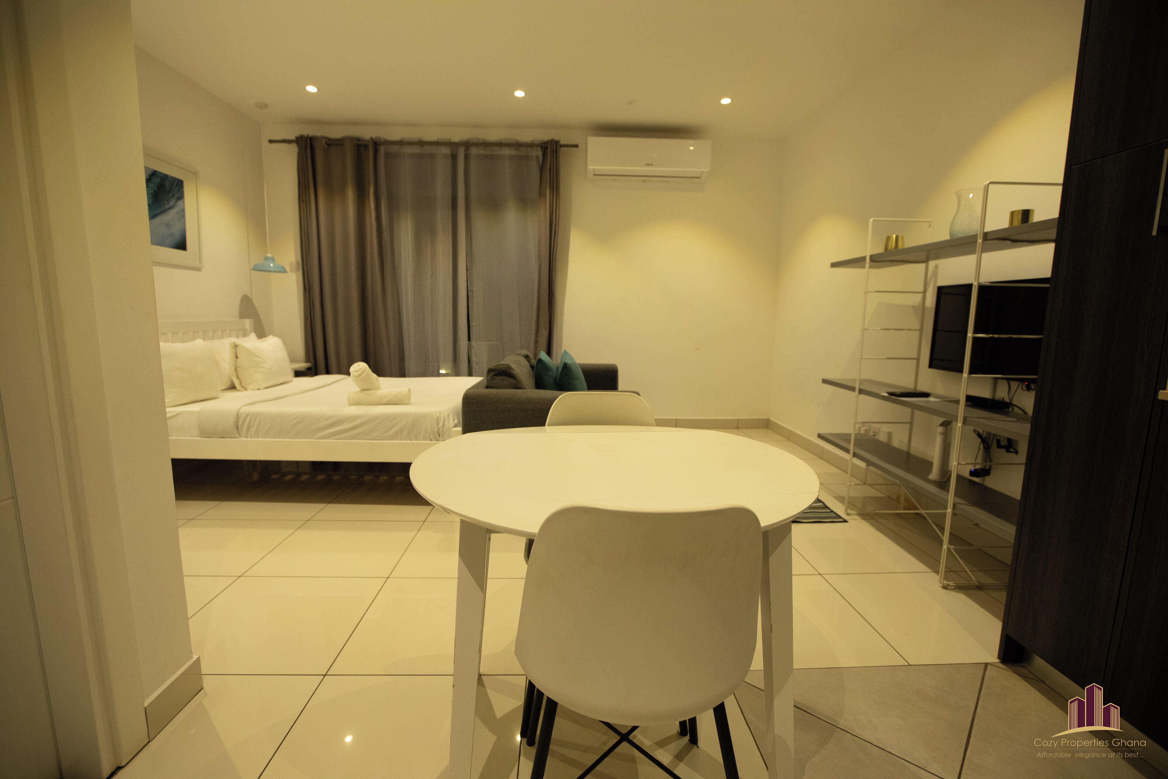 A modern hotel room with a bed, a dining table with a chair, a TV, and a built-in shelf with storage.