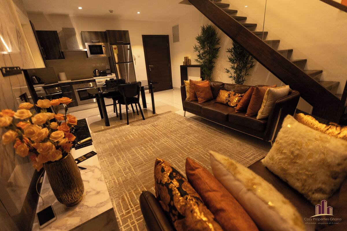 A modern kitchen with stainless steel appliances, a dining area with a table and chairs, a living room with a sofa and cushions, and a staircase leading to an upper level.