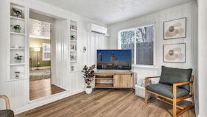 A living room with a wooden floor, a grey sofa, a wooden entertainment unit, and a large window with blinds.