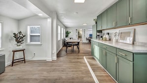 A modern kitchen with green cabinets, a dining area with a table and chairs, and a hallway with white walls and wooden flooring.