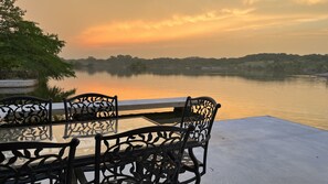 Une table en verre, entourée de chaises en métal aux motifs décoratifs, au bord d’un lac paisible, au coucher du soleil.