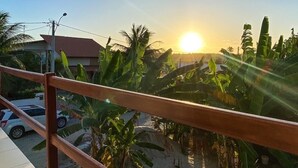 Varanda com vista para o pôr do sol, palmeiras e um edifício ao fundo.