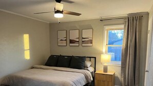 Quarto com cama, mesa de cabeceira, abajur, ventilador de teto e janela com cortinas.