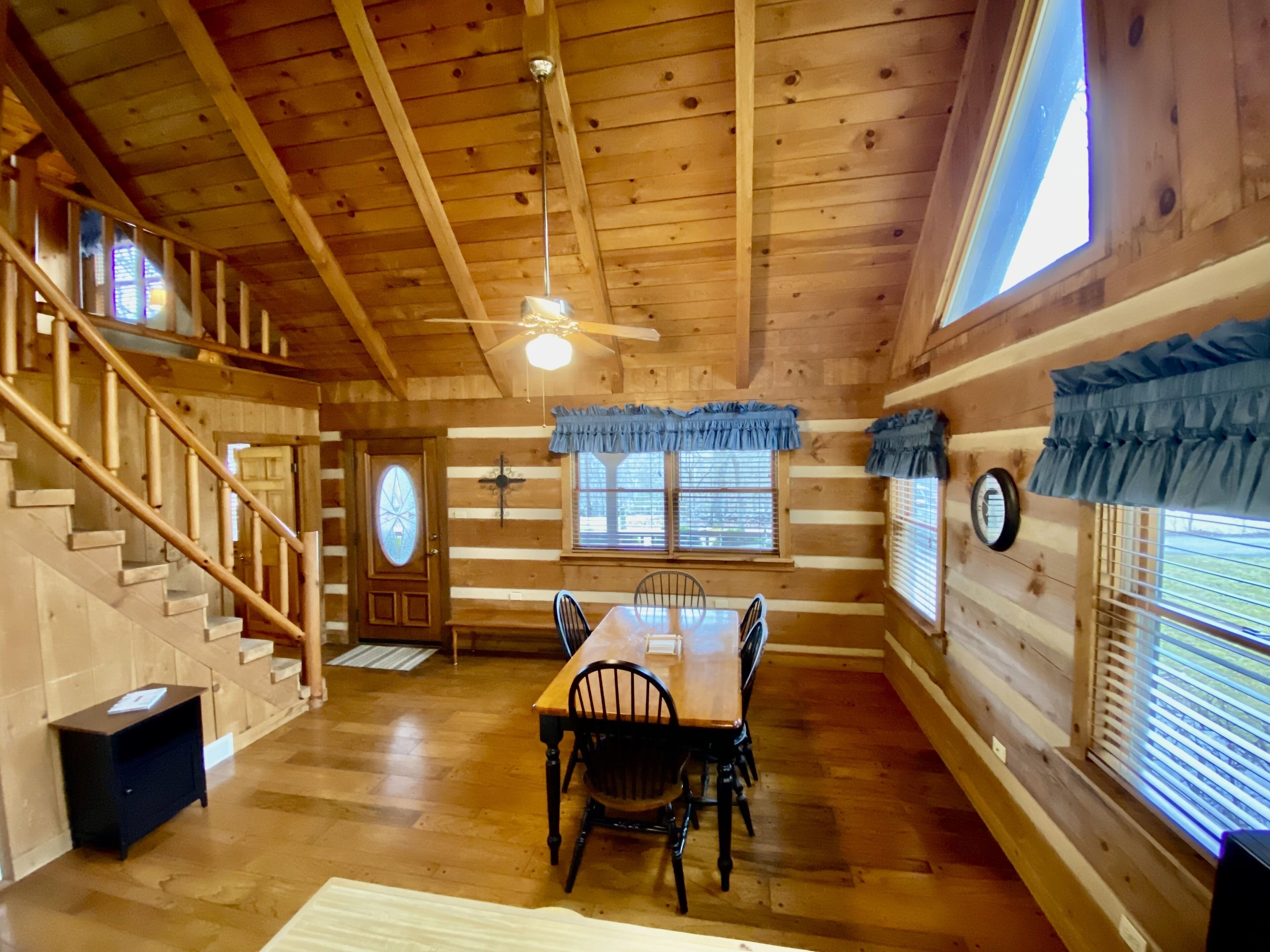 A wooden cabin interior with a dining area, staircase, and large windows.