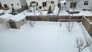 Un patio trasero cubierto de nieve, con una cerca de madera, un cobertizo pequeño y un árbol.
