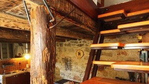 Un interior rústic amb bigues de fusta a la vista, una paret de pedra i una zona de cuina amb fogó i rentapesos.
