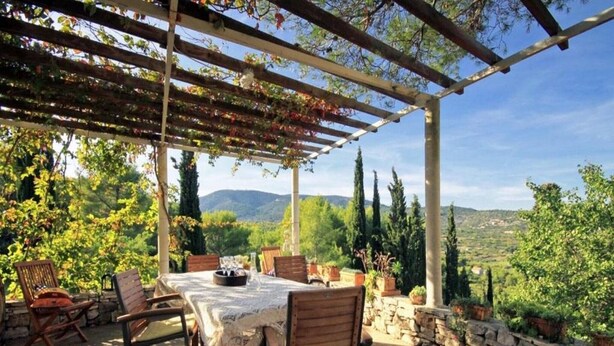 Terrassa exterior amb taula i cadires, envoltada de murs de pedra i plantes en testos, amb vista a un paisatge de muntanyes.