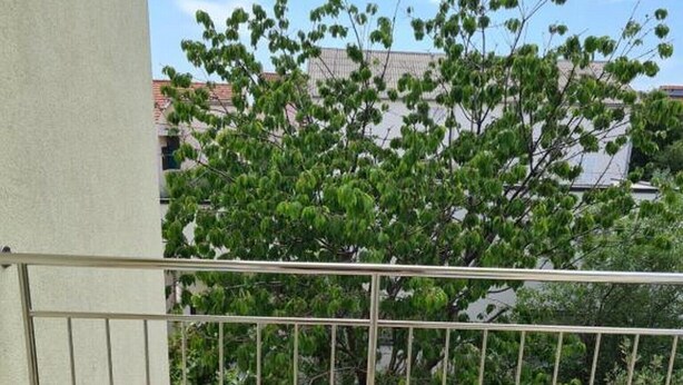 Ein Balkon mit Korbmöbeln und einem Glastisch, mit Blick auf Bäume und Gebäude.