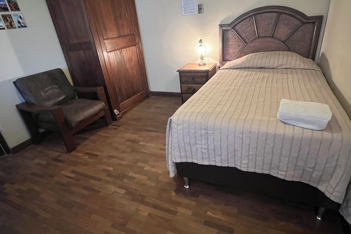 A bedroom with a wooden floor, a bed with a headboard, a nightstand with a lamp, a wooden wardrobe, and a chair.