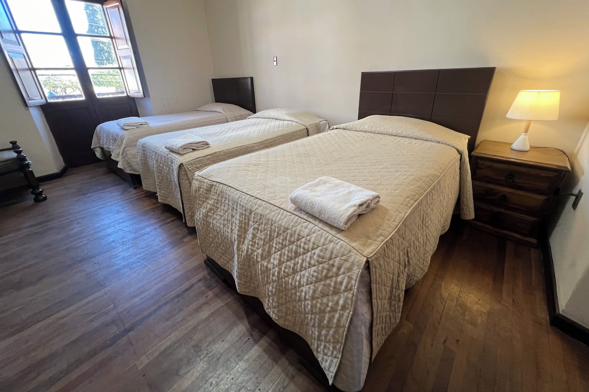 A hotel room with two beds, wooden flooring, a window, and a nightstand with a lamp.
