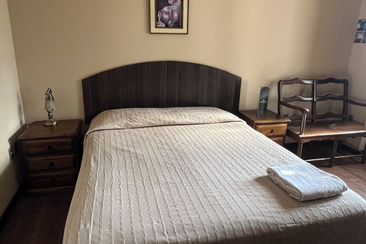 A neatly made bed with a quilted headboard, a bedside table with a lamp, a chair, and a framed picture on the wall.