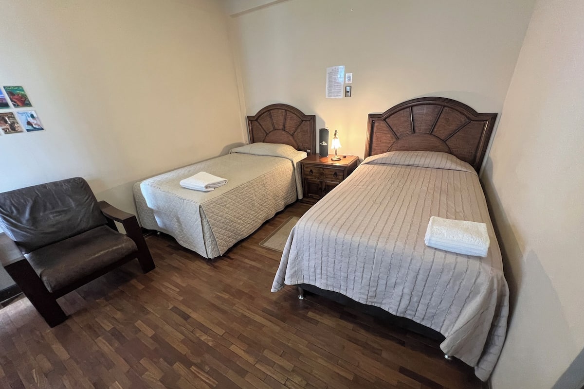 A hotel room with two single beds, a wooden floor, a chair, and a nightstand with a lamp.
