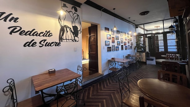 Interior de restaurante con una larga barra, mesas y sillas de madera. Se observa una pared con un mural y cuadros enmarcados.