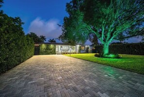 A house with a paved driveway leading to a front door, surrounded by well-maintained hedges and a large tree with illuminated green lights. - The Palms House Miami (Miami)