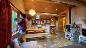 L’intérieur chaleureux d’un chalet avec un plafond en bois, une cheminée en pierre, un coin repas et une cuisine dotée de meubles blancs.