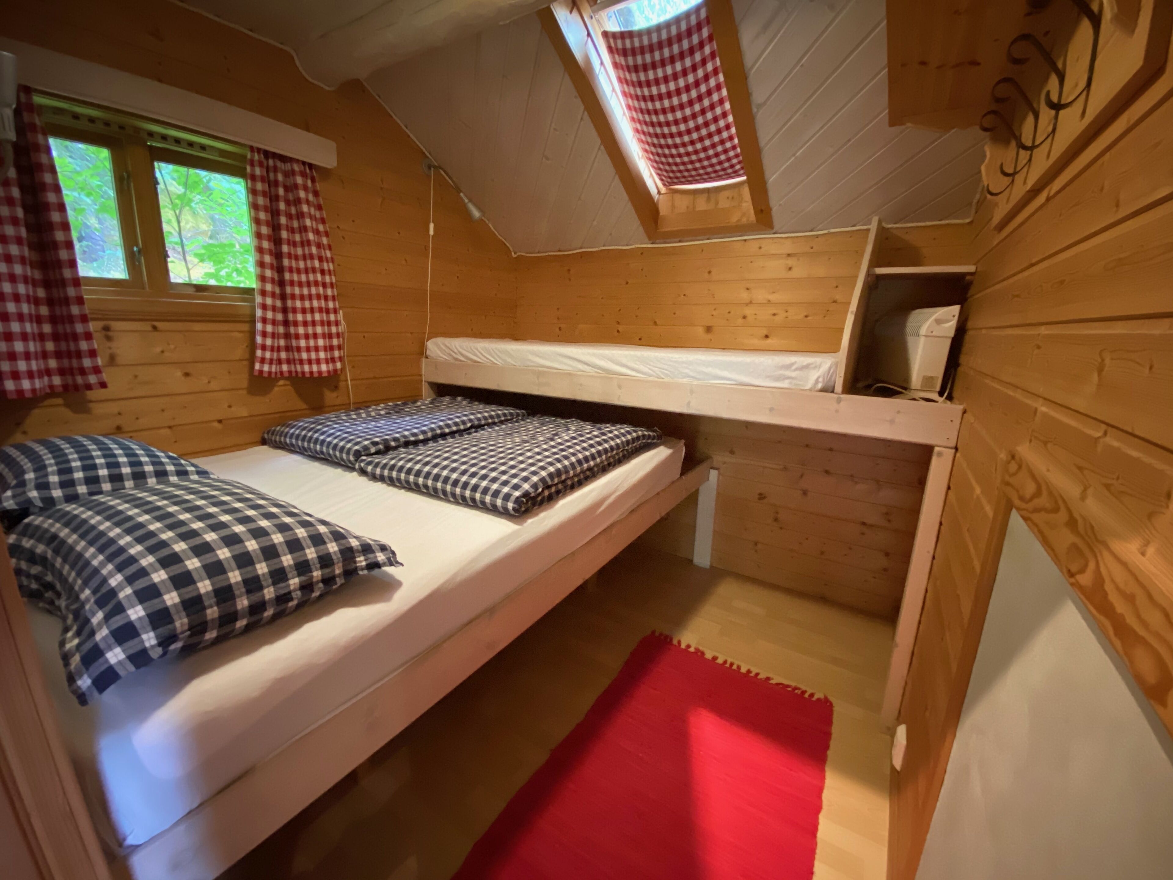 Una habitación pequeña y acogedora con paredes de madera y techo inclinado. Hay una cama nido con ropa de cama a cuadros y una alfombra roja en el suelo.