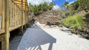 Une terrasse en bois agrémentée d’une pergola, de chaises longues blanches et d’une table avec des chaises, le tout entouré de galets blancs et de verdure.