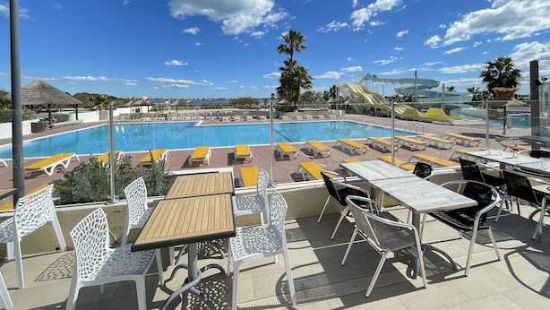 Un espace piscine sur le toit, avec des chaises longues et des tables, un ciel d’un bleu éclatant et un toboggan aquatique en arrière-plan.