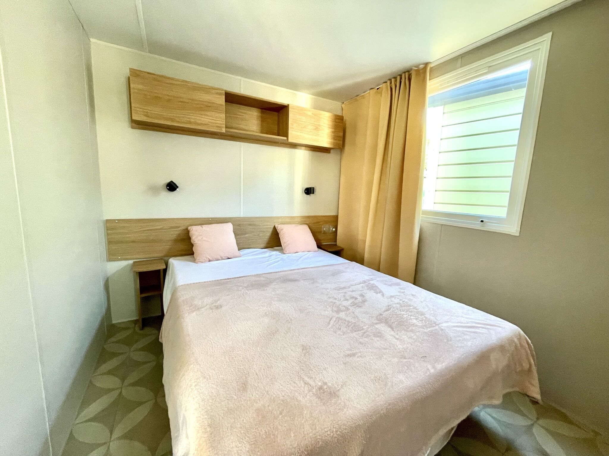 A bedroom with a bed, a wooden headboard, a window with curtains, and a wall-mounted shelf.