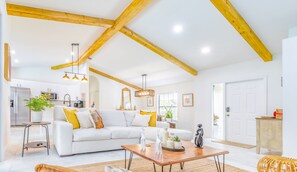 A spacious living room with a high ceiling and exposed wooden beams, a white sofa with yellow cushions, a wooden coffee table, and a kitchen area in the background.