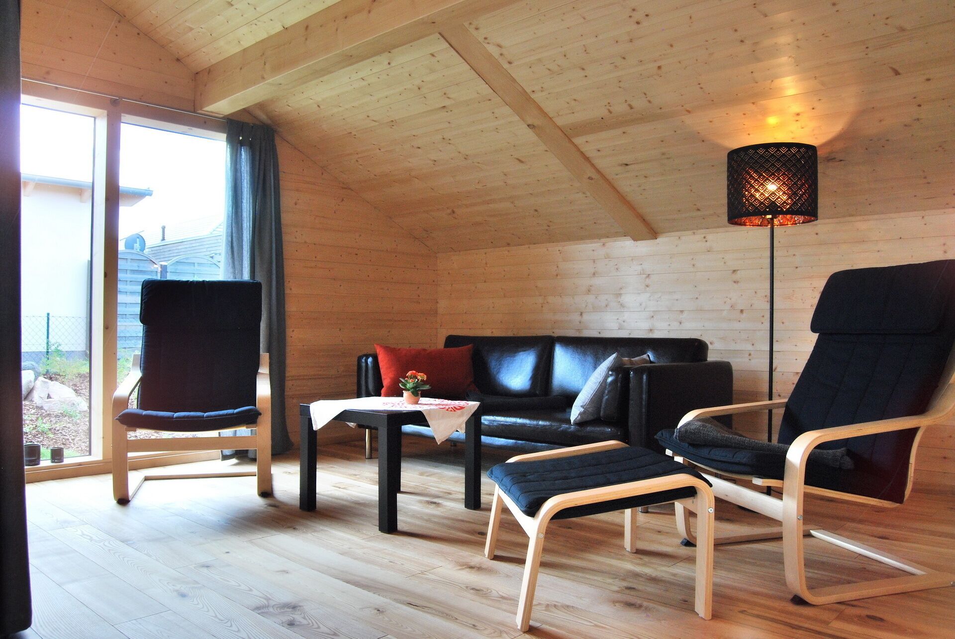 Una habitación acogedora con piso de madera, un sofá de cuero negro, dos sillones negros, una mesita y una lámpara de pie.