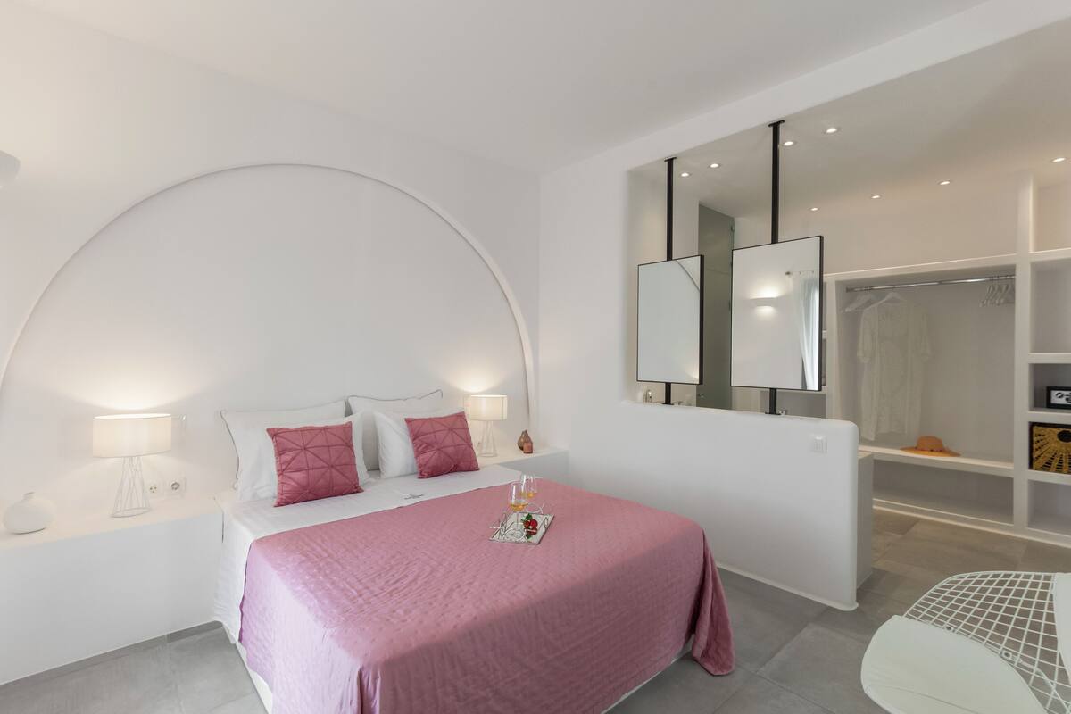 A modern bedroom with a curved ceiling, a large bed with pink bedding, bedside lamps, a mirror, and a walk-in closet.