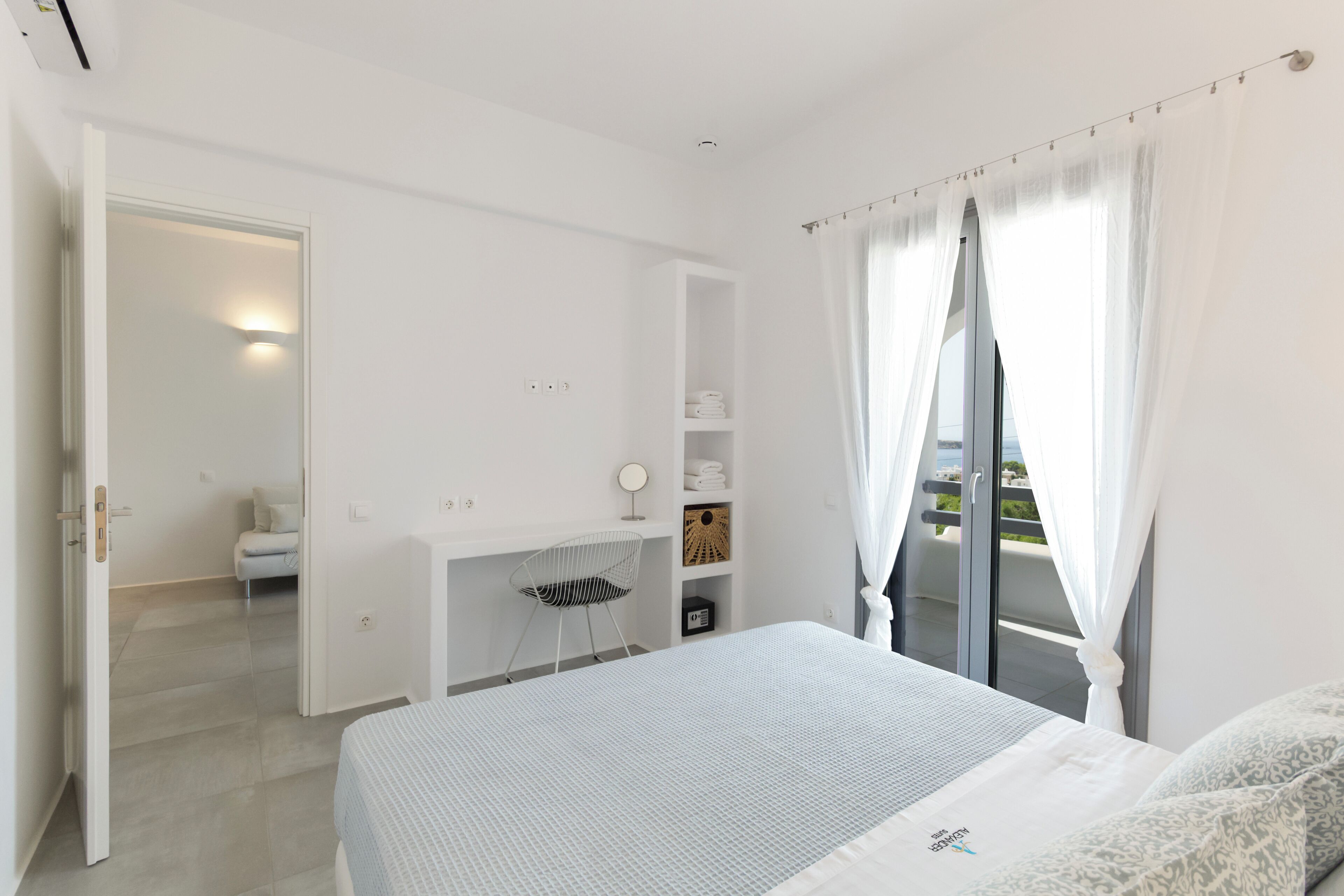 A bedroom with a bed, a desk, and a chair. There is a balcony with a view.