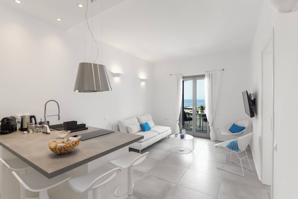 A modern kitchen with a dining area, white furniture, and a view of the sea through a glass door.