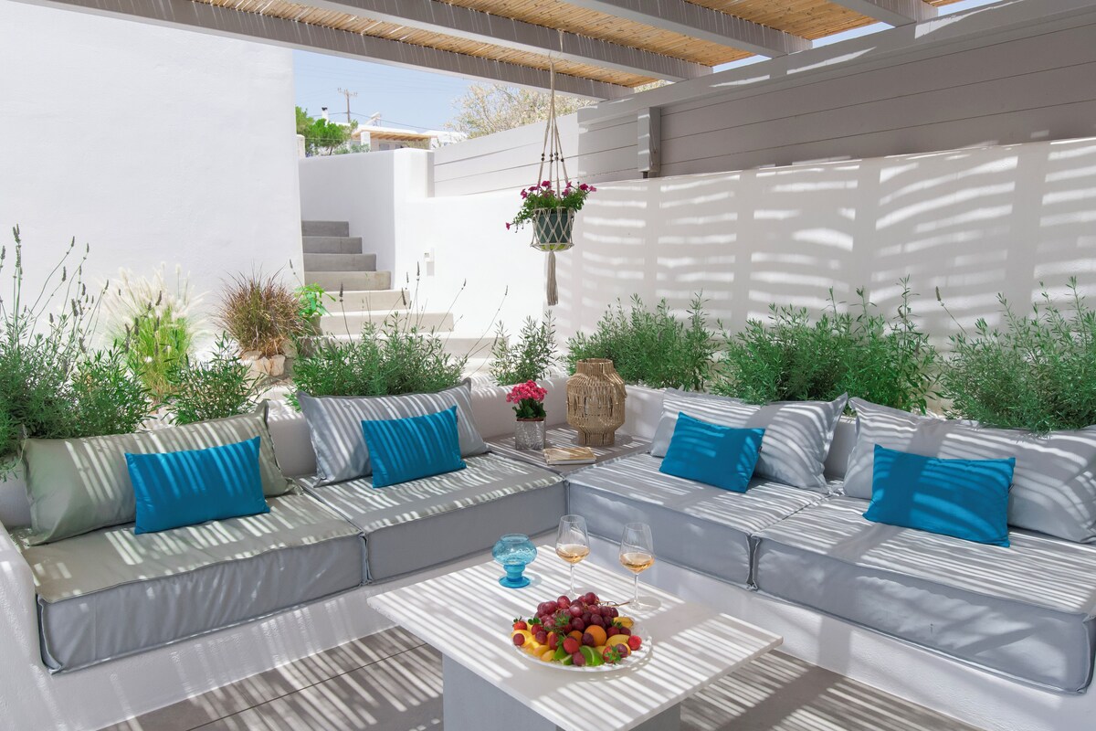 Ein moderner Außenbereich mit einer weißen Couch und blauen Kissen, einem Couchtisch mit Früchten und Gläsern sowie hängenden Pflanzen.