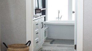 A bedroom with a bed, a bedside table, a dresser, a window, and a bathroom with a sink and mirror. - Cedar Ranch House Rental (Bandera)