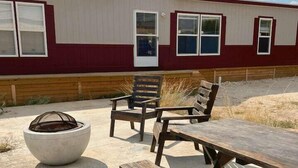 Un espace extérieur aménagé en terrasse, avec une table et des bancs en bois, un foyer et, au fond, un bâtiment à la façade rouge et blanche.
