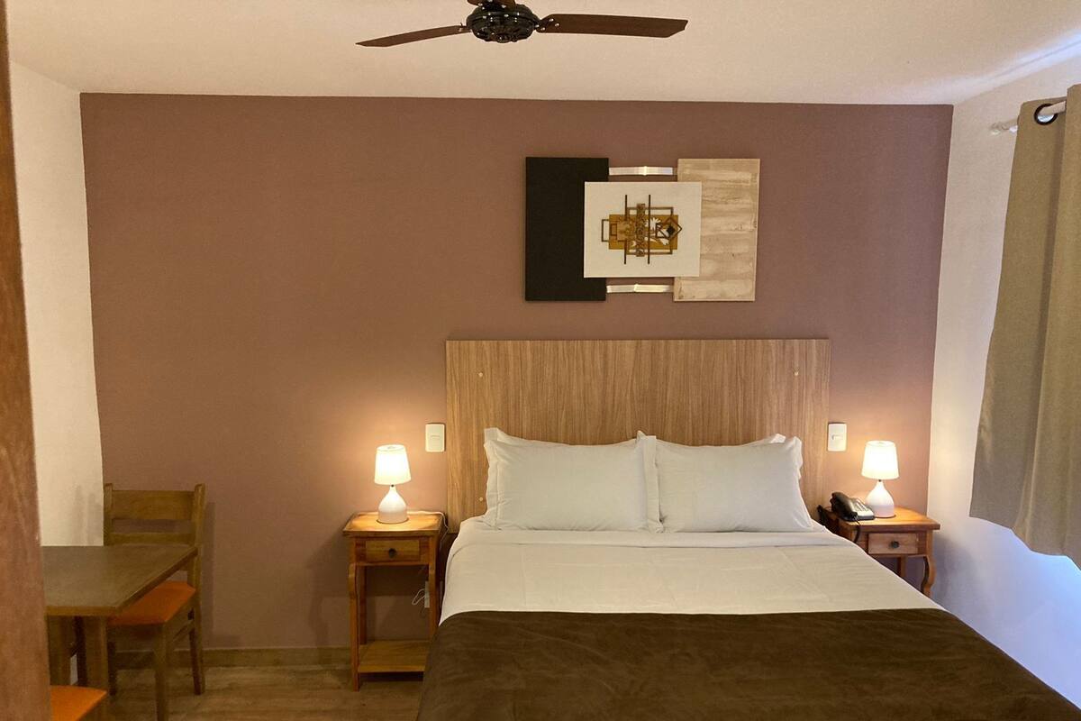 Une chambre d’hôtel comprenant un lit, deux tables de chevet avec des lampes, une tête de lit en bois, un ventilateur de plafond, une chaise et une œuvre d’art murale.