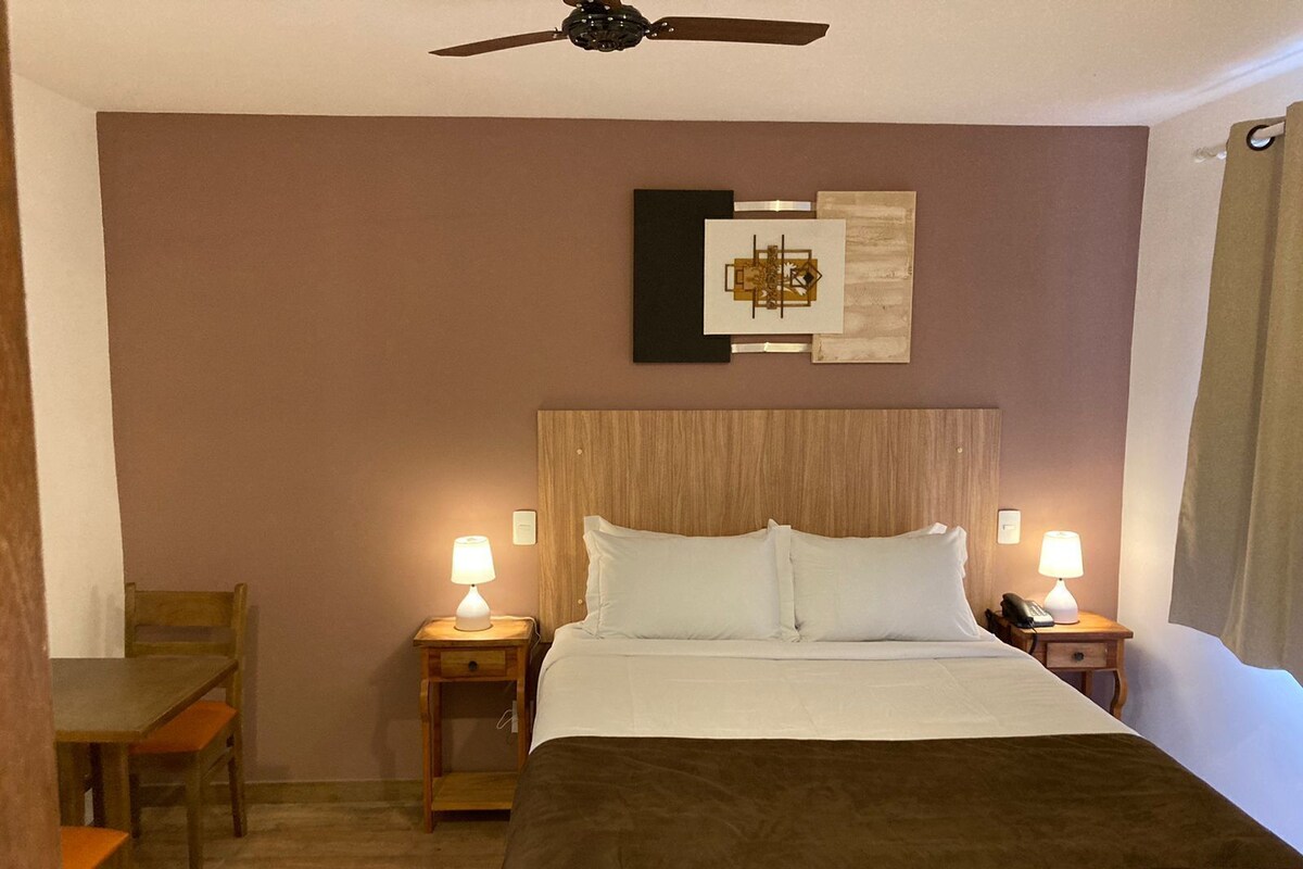 Une chambre d’hôtel comprenant un lit, deux tables de chevet avec des lampes, une chaise, un ventilateur de plafond et une œuvre d’art murale.