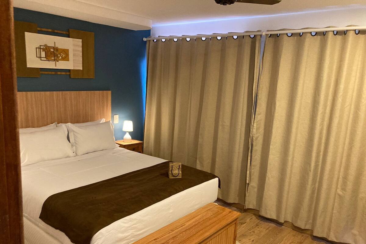 Un lit bien fait, avec une tête de lit en bois, une table de chevet avec une lampe et une fenêtre ornée de rideaux beiges.