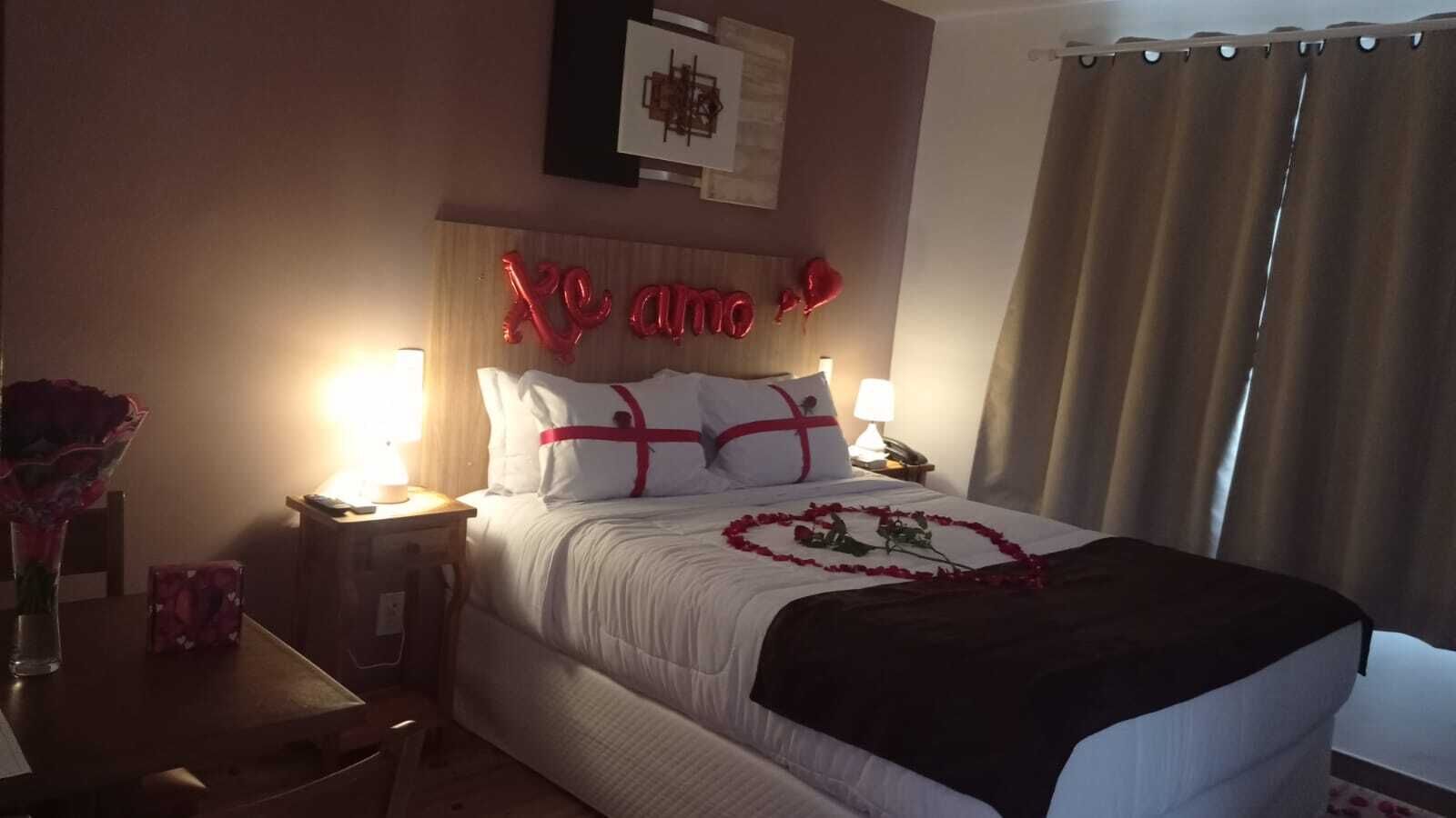 A hotel room with a bed, a bedside table with a lamp, a vase with flowers, and a decorative heart on the bed.
