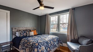 Un dormitorio con cama, ventilador de techo, ventana con cortinas y una silla.
