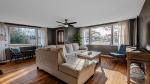 Un salón amplio con un sofá seccional color beige, una mesa de centro y un mueble para el televisor. Hay ventanas grandes que ofrecen vista a árboles y un edificio.