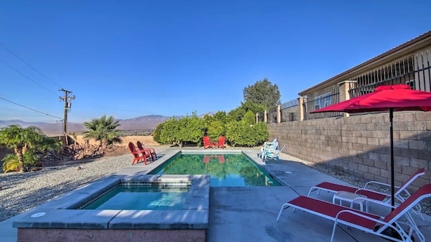 A swimming pool with red chairs and a red umbrella, surrounded by a stone wall and greenery.