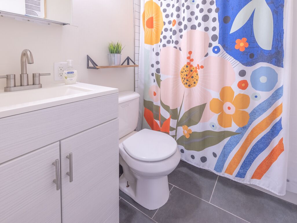Ein Badezimmer mit weißem Waschbecken, Wasserhahn und Toilette. Ein Duschvorhang mit floralem Muster ist angebracht.