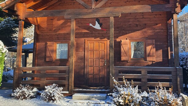 Une cabane en bois, dont le toit est recouvert de neige, avec une porte centrale et deux fenêtres de chaque côté.
