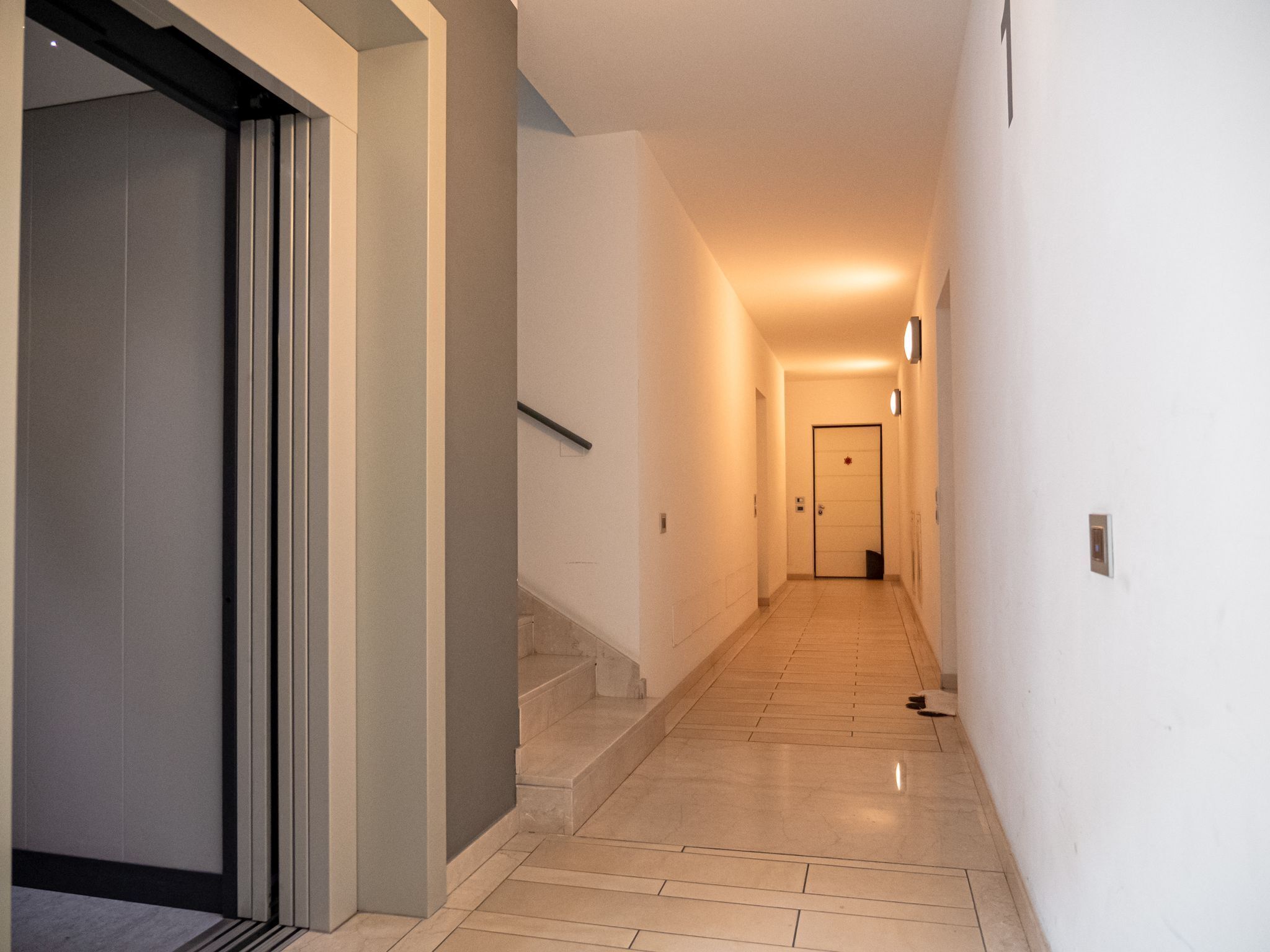 Un pasillo con una puerta al final, una escalera y una lámpara en la pared.