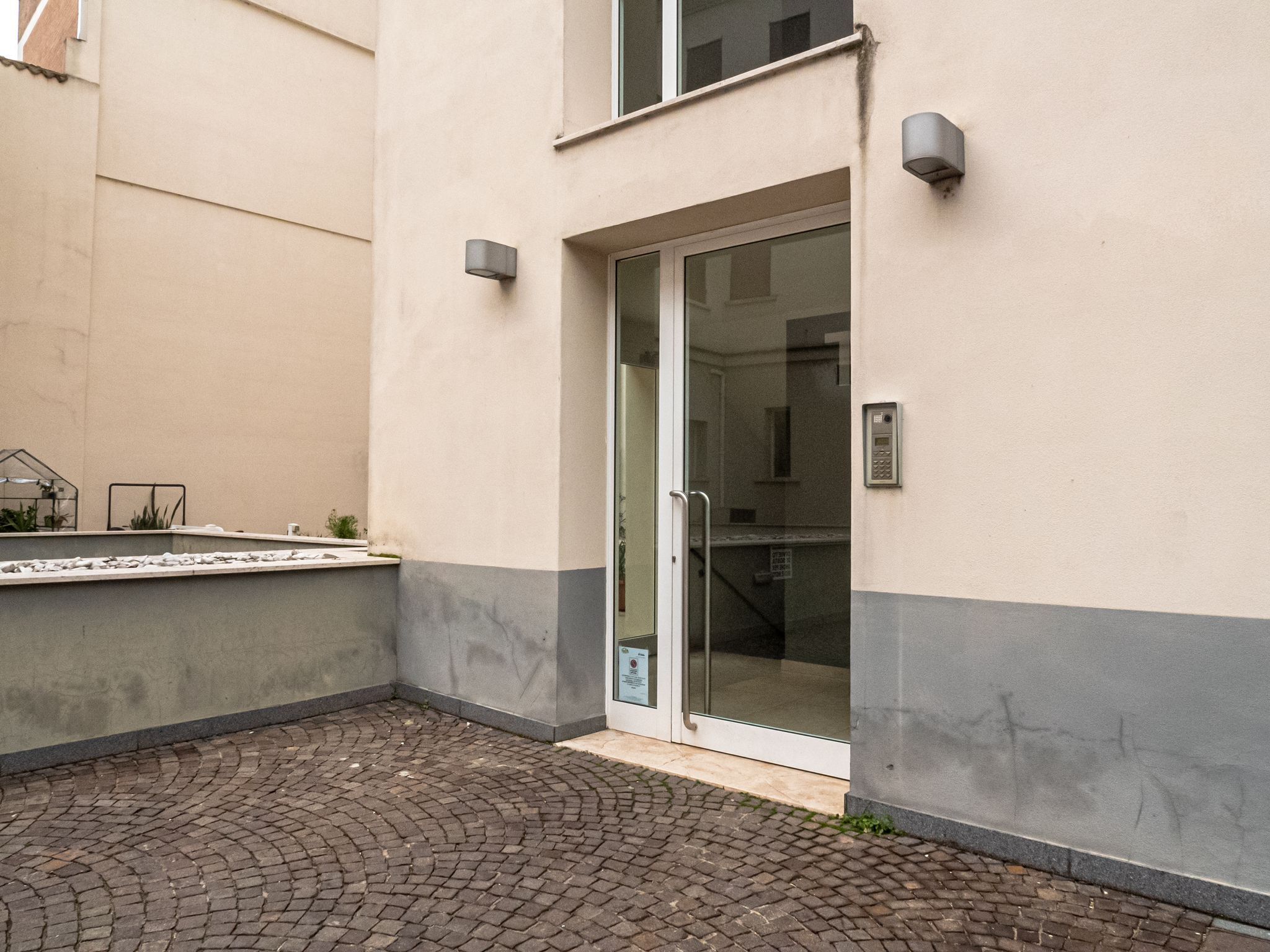 A building with a glass door entrance, a small outdoor seating area with plants, and a paved courtyard.