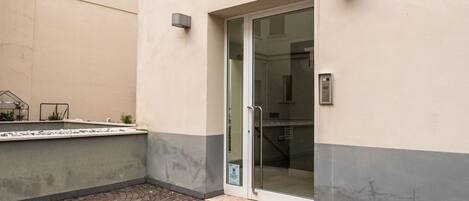 A building with a glass door entrance, a small outdoor seating area with plants, and a paved courtyard.