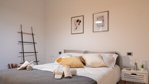 Una cama bien hecha con almohadas y una manta, una mesita de noche con una lámpara y una planta, y dos cuadros en la pared.